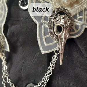 May include: A silver skull brooch with a chain attached. The brooch is on a black fabric background. The skull is detailed and has a bird-like beak.