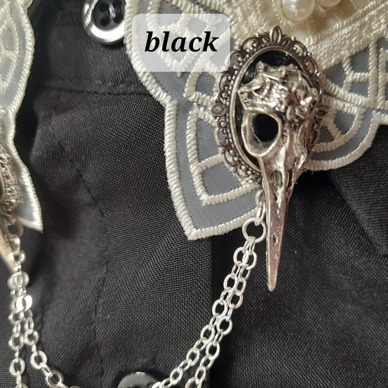 May include: A silver skull brooch with a chain attached. The brooch is on a black fabric background. The skull is detailed and has a bird-like beak.