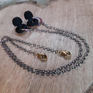 May include: A black earbud holder with a chain. The chain is a dark, iridescent color with gold-colored clasps. The earbud holders are black and have a circular design. The background is a wooden surface.
