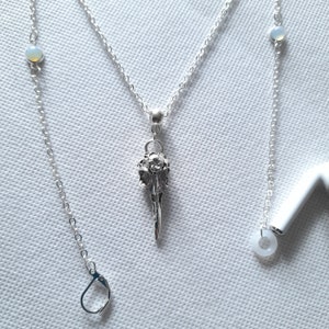May include: A silver chain necklace with a bird skull pendant and two opal beads. The necklace is on a white background.