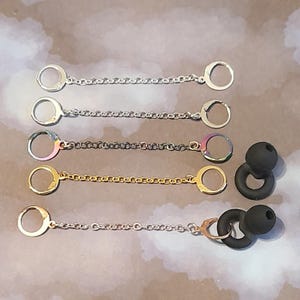 May include: Earbud accessories featuring four metal chain designs with circular clasps in silver, iridescent, gold, and silver. Two black earbud tips are also included, with a ring-shaped holder.
