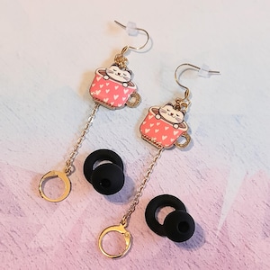May include: A pair of dangle earrings featuring a pink teacup design with white hearts and a cat peeking over the rim. The earrings have gold-colored hooks and chains, with black earbud tips attached.