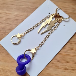 May include: Gold-colored earrings with a lightning bolt and star design. The earrings have a chain and a purple earplug-shaped charm. The earrings are displayed on a white card.