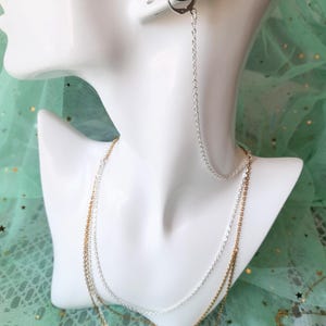 May include: A silver-toned chain earring connector that attaches to a black ear cuff. The long, delicate silver chain drapes down to connect with a layered necklace featuring both silver and gold-toned metal chains on a white display bust.