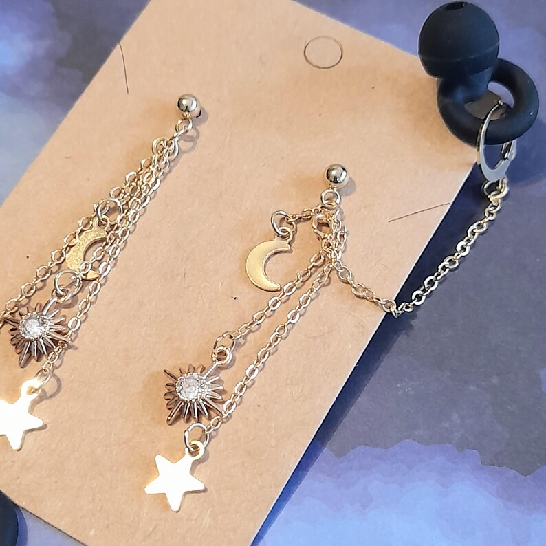 Puede incluir: Pendientes dorados con tem&aacute;tica celestial. Un pendiente presenta un dise&ntilde;o de sol, luna y estrella con cadenas colgantes. El otro pendiente tiene un dise&ntilde;o de sol, luna y estrella con cadenas colgantes. Los pendientes est&aacute;n conectados por una cadena y un cierre de goma negro.