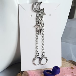 May include: Silver-coloured dangle earrings with a celestial theme. The earrings feature a hoop design with small clear stones, a star-shaped charm, and dangling chains ending in small hoops. The earrings are displayed on a white card.