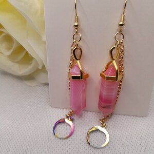 May include: A pair of gold dangle earrings with pink and white agate stone pendants. The pendants are shaped like a long, thin point and are attached to a gold chain with a small gold hoop at the bottom.