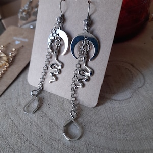 May include: Silver dangle earrings with a crescent moon and snake charm. The earrings have a drop design and are attached to a hook.