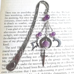 May include: Silver metal bookmark with a sun design and decorative charms. Charms include a star, crescent moon, and a dagger, with purple amethyst beads. The bookmark is on a book page.