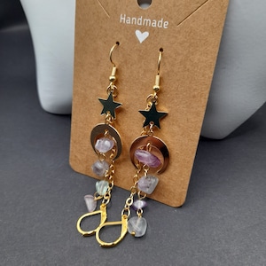 May include: Gold-coloured drop earrings with star and crescent moon charms, and hanging, irregularly shaped, light purple and green gemstones. The earrings are displayed on a brown card with the word "Handmade" and a heart.