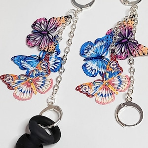 May include: A pair of earplug accessories featuring silver-tone metal chains and multiple colorful butterfly charms in shades of blue, purple, orange, and yellow. Each chain connects to a silver hoop and a black silicone earplug attachment.