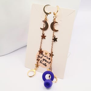 May include: Gold earrings with a crescent moon and star charms hanging from a chain. The earrings have a purple silicone plug on the bottom.