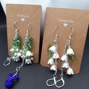 May include: Two pairs of handmade earrings featuring white lily of the valley flowers and green leaves. The earrings are on brown cards with the word "Handmade" written on them.