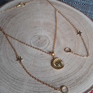 May include: A gold-colored necklace with a sun and moon pendant. The necklace has a delicate chain design with star and ring accents. The jewelry is displayed on a wooden surface.