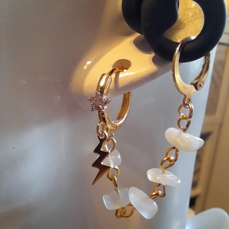 May include: A gold-toned hoop earring featuring a small star charm with clear stones, a gold lightning bolt charm, and a dangling chain segment adorned with several smooth, white, irregular-shaped stone beads that connect to the main hoop.