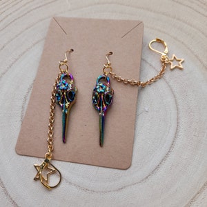 May include: A pair of iridescent bird skull earrings with gold-colored chains and ear hooks. One earring features a dangling star charm. The earrings are displayed on a brown card against a wooden background.