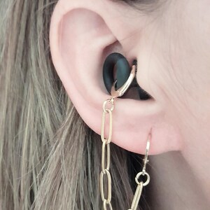 May include: A gold-toned metal chain earring featuring a series of elongated oval links. The chain connects a small hoop earring inserted into the conch area of the ear to a second hoop earring positioned in the earlobe, encircling a black ear plug.