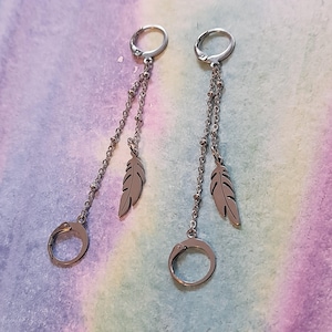 May include: A pair of silver-toned dangle earrings. Each earring features a small hoop at the top, a beaded chain, a feather charm, and a larger hoop at the bottom. The earrings are displayed against a pastel-colored background.
