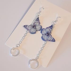 May include: A pair of silver dangle earrings featuring a butterfly design. The butterflies are a light blue and purple colour with a silver chain hanging from each butterfly.