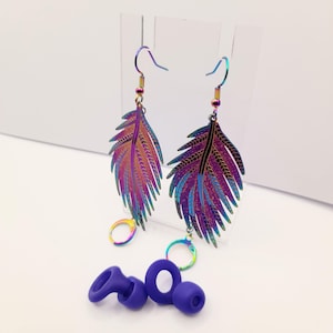 May include: A pair of iridescent feather earrings with a rainbow color scheme. The earrings are made of metal and have a hook closure. The earrings are shown on a white background with a pair of purple earplugs.
