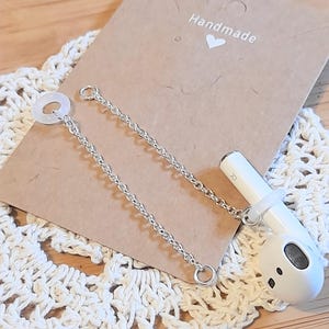 May include: A silver-toned metal chain earring attachment designed for a white wireless earbud. The accessory features a thin link chain connected to a small clear silicone ring that slides onto the earbud stem, displayed on a brown card with the text Handmade and a heart icon.