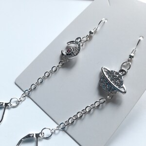 May include: A pair of silver earrings featuring a crescent moon and a small planet charm. The charms are decorated with clear rhinestones and a pink stone in the centre of the moon.