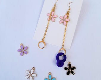 May include: Dangle earrings featuring pink flower charms and gold chains. Additional flower charms in purple, white, black, and blue are scattered around. The earrings have a gold ring and a dark blue ring.