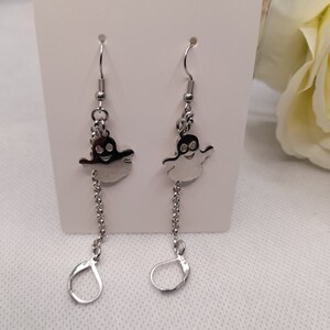 May include: A pair of silver dangle earrings featuring a ghost charm with a smiling face. The ghost charm is attached to a silver chain that hangs from the earring hook.