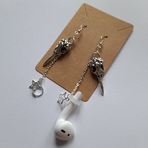 May include: Silver earrings with bird skull pendants, chain details, and star charms. One earring is styled with a white earbud. The earrings are presented on a brown card against a white backdrop.