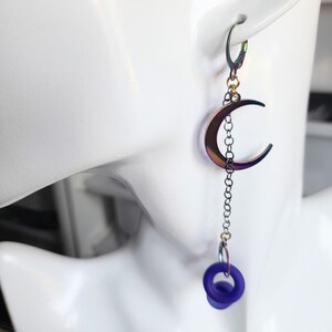 May include: A single, iridescent crescent moon earring with a chain and a dangling dark blue ring. The earring is attached to a silver-coloured lever-back closure. The moon has a metallic, colour-shifting finish.