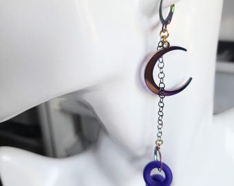May include: A single, iridescent crescent moon earring with a chain and a dangling dark blue ring. The earring is attached to a silver-coloured lever-back closure. The moon has a metallic, colour-shifting finish.