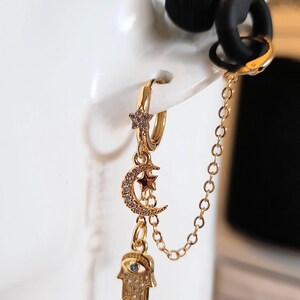 May include: A gold-toned double hoop earring connected by a delicate chain. The main hoop features a small star charm, while a crescent moon charm and a hamsa hand charm with a blue centre stone hang below. The earring is worn with a black earbud.