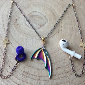 May include: A rainbow iridescent chain with a pair of star charms, a pair of earbud holders, and a single wing charm. The earbud holders are purple and the wing charm is iridescent.