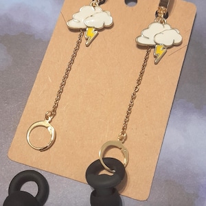 May include: Gold-tone earrings featuring white cloud charms with yellow lightning bolts. The earrings have a long chain and a circular clasp. Two black earbud tips are also visible.