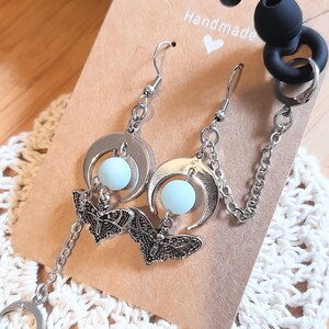 May include: Silver-toned earrings featuring a crescent moon charm, a light blue spherical bead, and a detailed moth pendant. The earrings include a silver chain attachment with a black earbud cover and a small metal hoop, all displayed on a brown card.