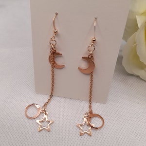 May include: A pair of rose gold dangle earrings with a crescent moon charm, a star charm, and a circle charm hanging from a chain.