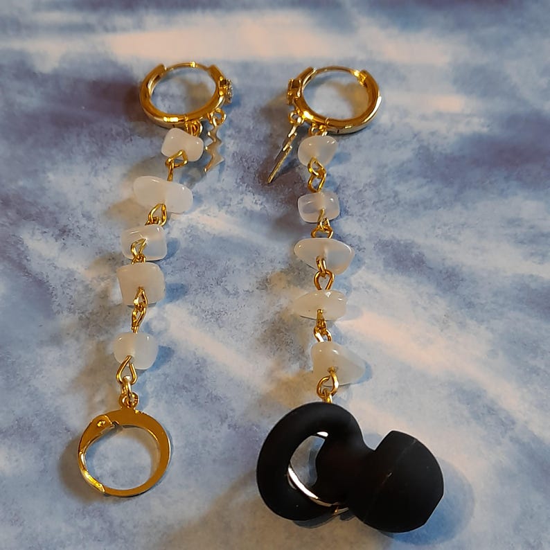 May include: A pair of gold-tone hoop earrings featuring dangling chains of white, irregular, tumbled stone beads. One earring ends in a gold-tone hoop, while the other earring is attached to a black, circular silicone earbud cover.
