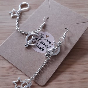 May include: Silver dangle earrings with a cat sitting on a crescent moon charm and a star charm hanging from a chain. The earrings are on a brown card with the text "You've got great taste".