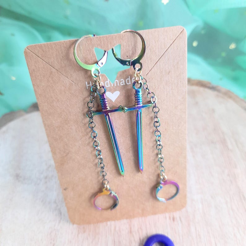 May include: A pair of iridescent sword earrings with a chain and handcuff charm. The earrings are attached to a brown card with the word "Handmade" and a heart symbol. The earrings are displayed on a wooden surface.