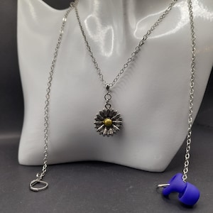 May include: A silver chain necklace with a daisy pendant. The daisy has a gold centre and silver petals. The necklace is on a white display stand. There is a blue earbud on the right side of the image.