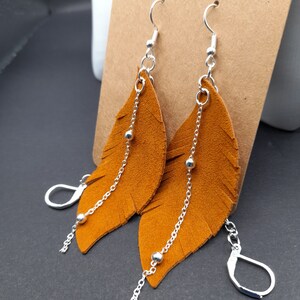 May include: A pair of dangle earrings featuring a brown suede feather design with silver chain accents. Each earring has three silver beads on the chain.