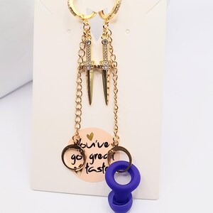 May include: Gold earrings with a dangling dagger charm and a small blue silicone charm. The earrings are on a white background with the text "You've got great taste".