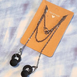 Peut inclure: Un collier chaîne argenté avec des supports d'écouteurs noirs. Le collier est attaché à une carte marron avec l'inscription "Handmade" et un petit cœur. Le fond est un motif marbré bleu clair et blanc.
