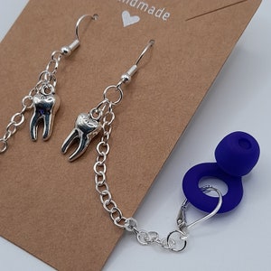 May include: Silver tooth charm earrings with a silver chain and blue silicone earplugs. The earrings are on a brown card with the word "handmade" written on it.