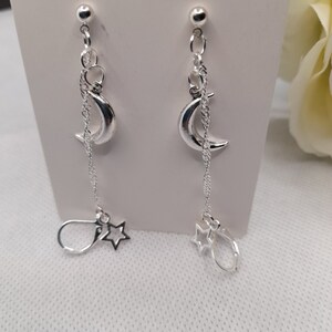 May include: Silver dangle earrings with a crescent moon charm and a star charm hanging from a chain. The earrings are on a white background.