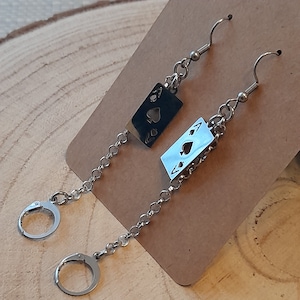 May include: A pair of silver earrings with a dangling card suit charm. Each charm features a cut-out ace of spades design.