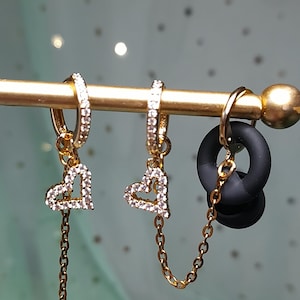 May include: Gold-toned earrings with heart-shaped pendants encrusted with clear crystals. One pair features a dangling chain. A black, circular plug is also visible, connected by a chain to one earring. The jewelry is displayed on a gold bar.