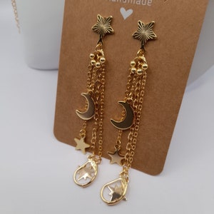 May include: Gold-coloured celestial earrings with star and crescent moon charms. The earrings have a star-shaped top, multiple chain strands, and dangling charms. The earrings are displayed on a brown card.