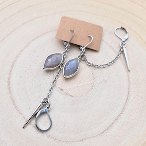 May include: A pair of silver earrings with a light gray stone. The earrings are connected by a silver chain with a silver hook on one end.