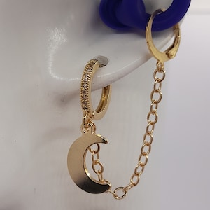 May include: Gold hoop earring with a crescent moon charm and a chain dangling from the bottom. The earring is set with small clear stones.